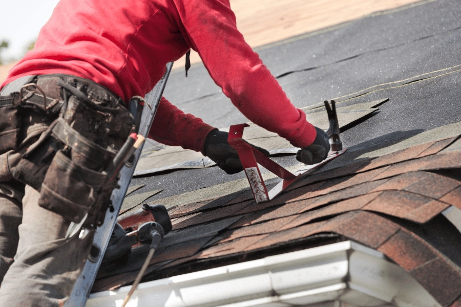 worker repairing a home roof standing on a ladder with a hammer